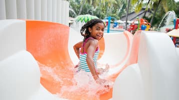 Un niño resbalando por un tobogán acuático con diseño blanco y naranja, rodeado de palmeras y una zona de piscina.