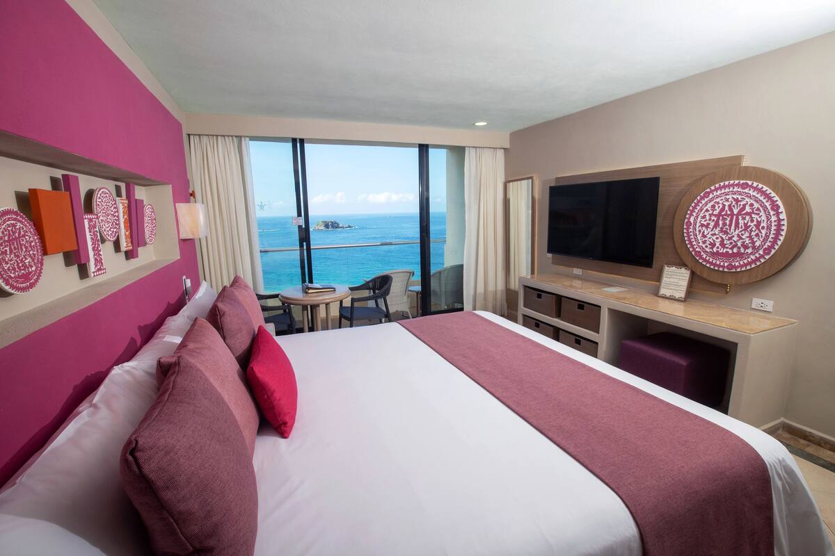 A hotel room with a large bed, a desk with a flat-screen TV, and a view of the ocean.