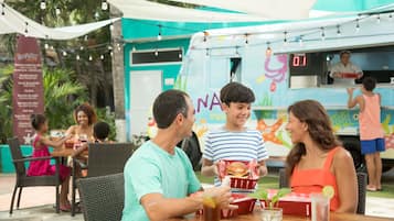 An outdoor dining area with wicker chairs and tables, a food truck with colorful branding, and a tropical backdrop.