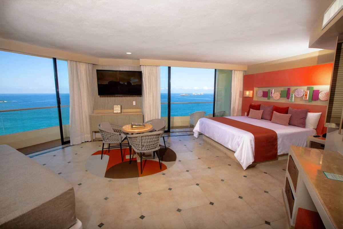 A hotel room with a bed, a TV, a dining table, and a view of the ocean.