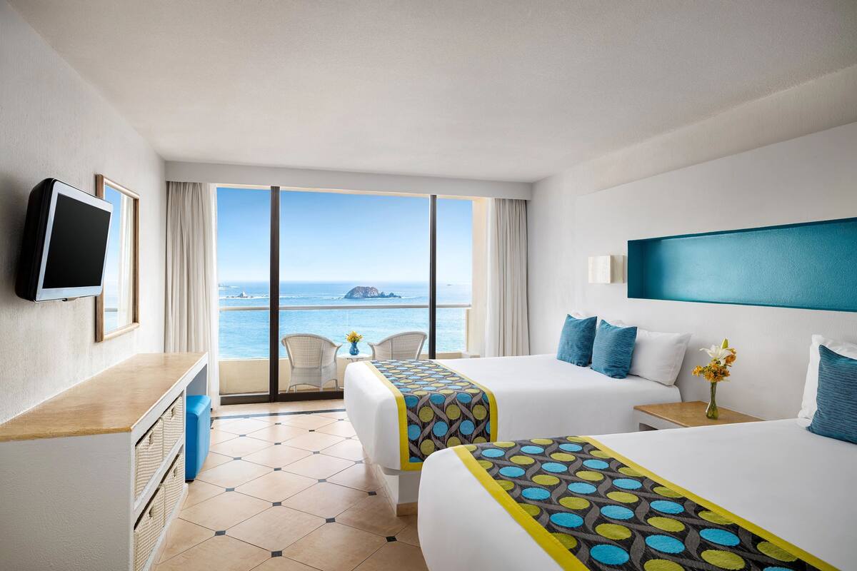 A hotel room with two beds, a TV, a desk, and a view of the sea.