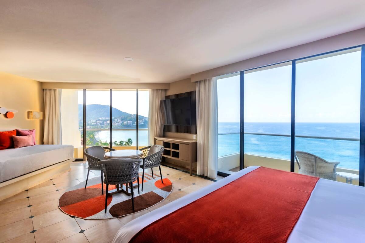 A hotel room with a bed, a desk, and a balcony with a view of the ocean.