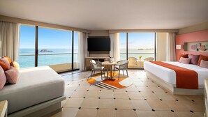 A hotel room with a large bed, a sofa, a dining table with chairs, a TV, and a view of the sea.