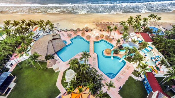 An aerial view of a resort with two large swimming pools, a beach, and palm trees.