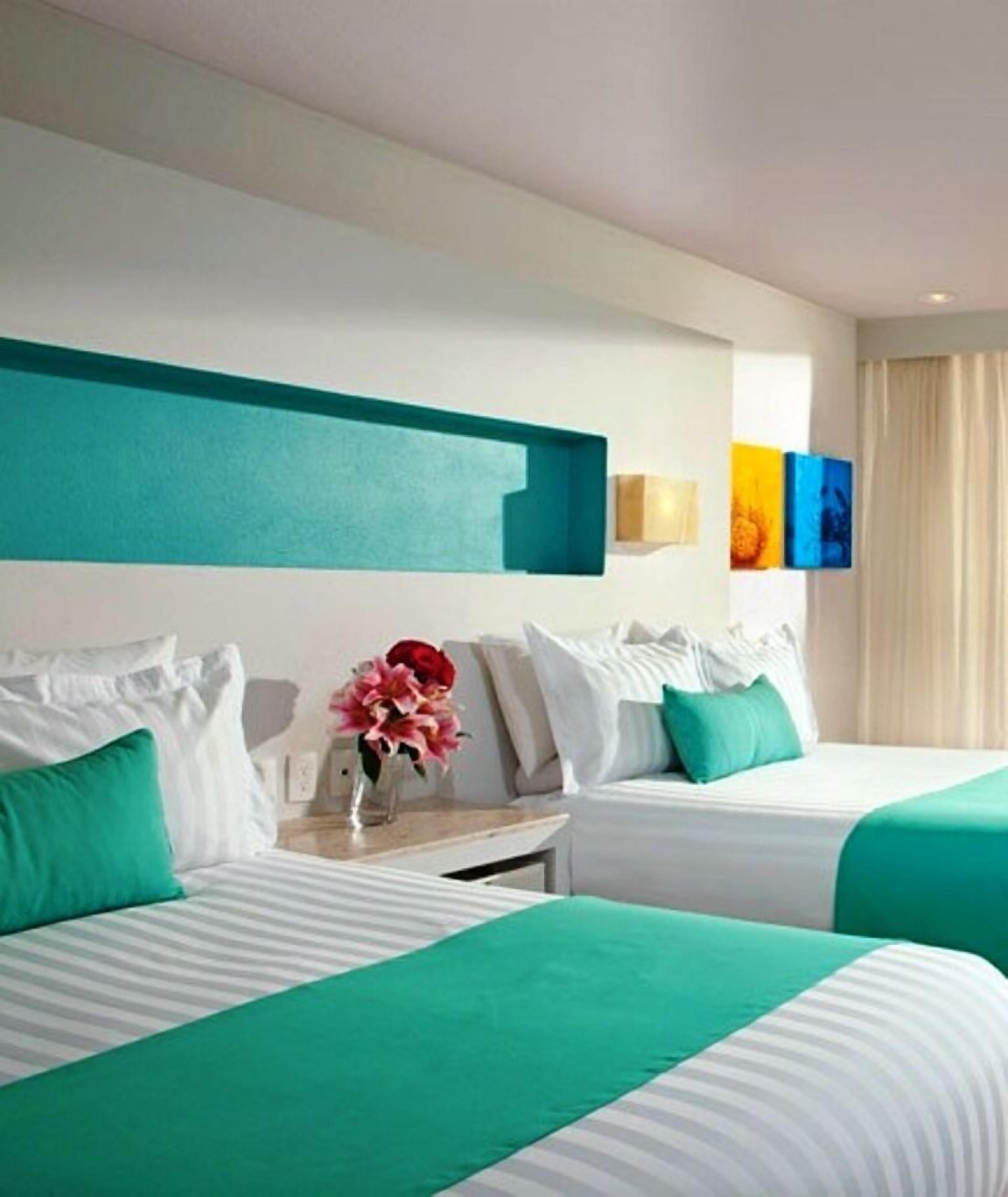 Una habitación de hotel con dos camas, ropa de cama color aguamarina, almohadas blancas, una mesita de noche con un jarrón de flores y una pared con detalles en aguamarina y obras de arte coloridas.