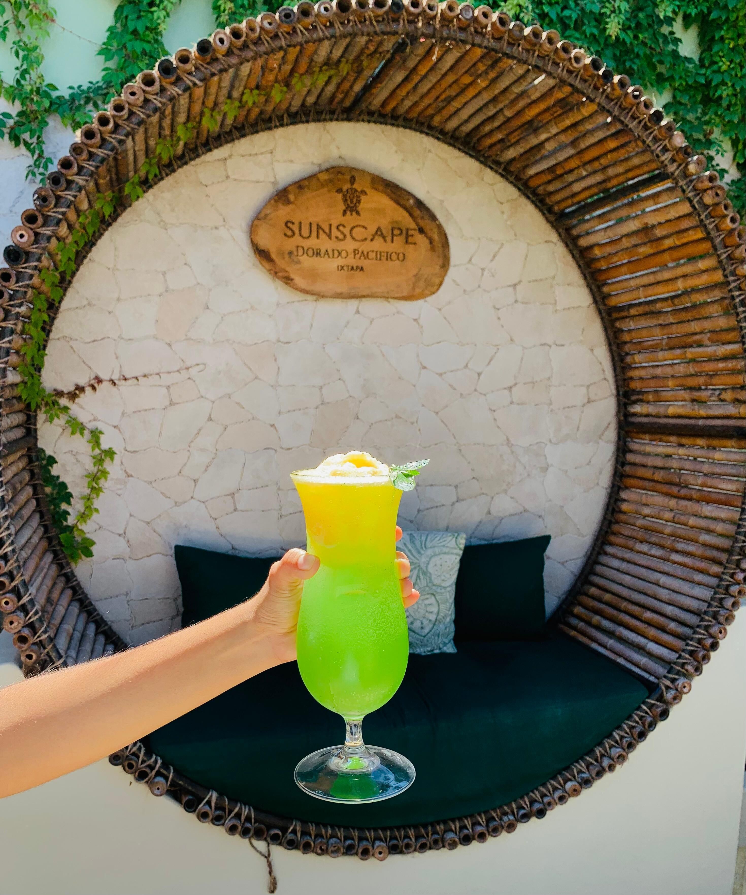 A hand holding a green cocktail with a garnish, in front of a circular wicker frame with a sign reading "SUNSCAPE DORADO PACIFICO IXTAPA".