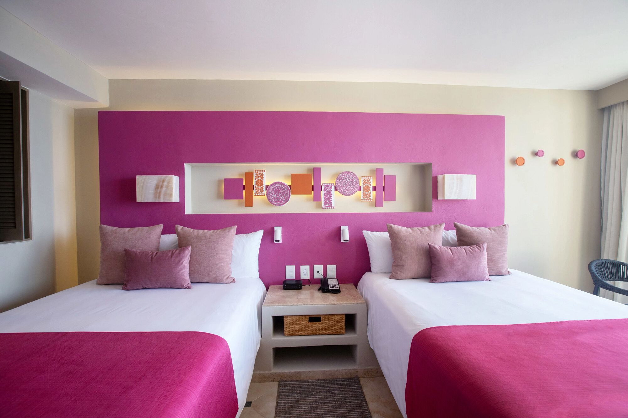 Habitación de hotel con dos camas, cabeceros con paneles decorativos, mesitas de noche con lámparas y una pequeña zona de estar con una silla.