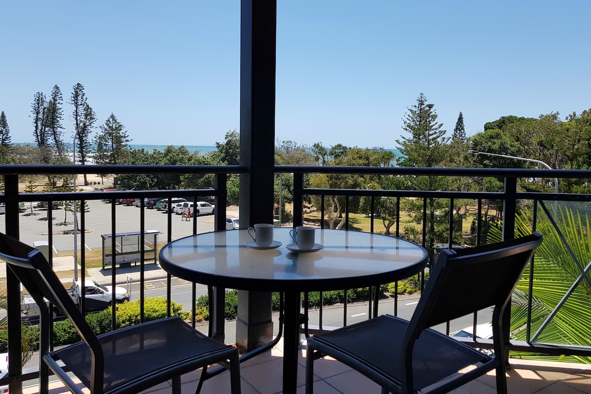 A balcony with a table and chairs, overlooking a parking area and a view of trees and a body of water.