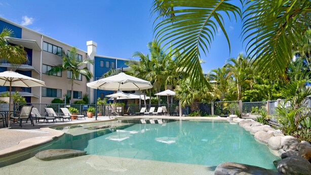 Zona de piscina con sillas de descanso y sombrillas, rodeada de palmeras y un edificio con balcones.