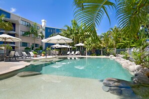 A pool area with lounge chairs and umbrellas, surrounded by palm trees and a building with balconies.