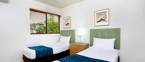 Una camera d'albergo con due letti, una ventola a soffitto, una finestra con tende, un comodino con una lampada e quadri appesi al muro.