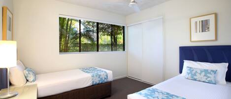 A hotel room with two beds, a ceiling fan, a window with blinds, a framed picture on the wall, and a nightstand with a lamp.