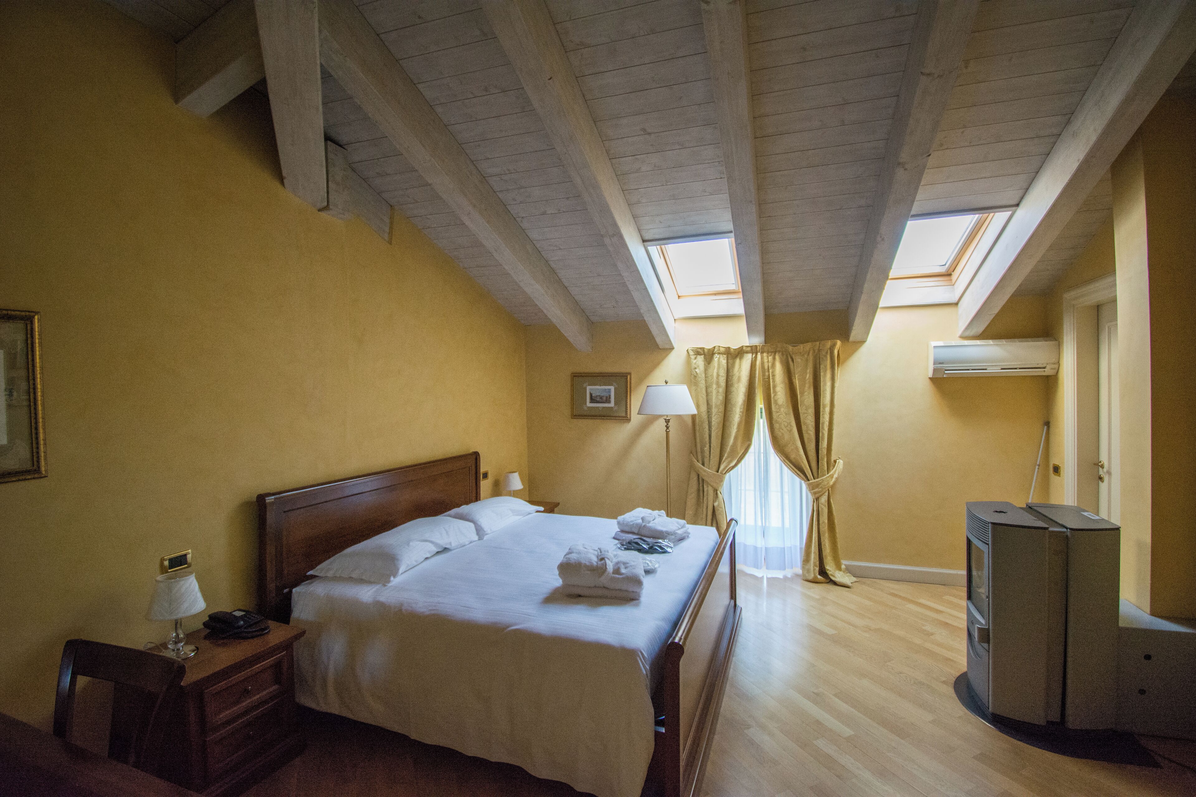 Habitación de hotel con una cama de madera, una mesita de noche con lámpara, un escritorio con teléfono y un calentador. Hay un tragaluz sobre la cama y una ventana con cortinas.