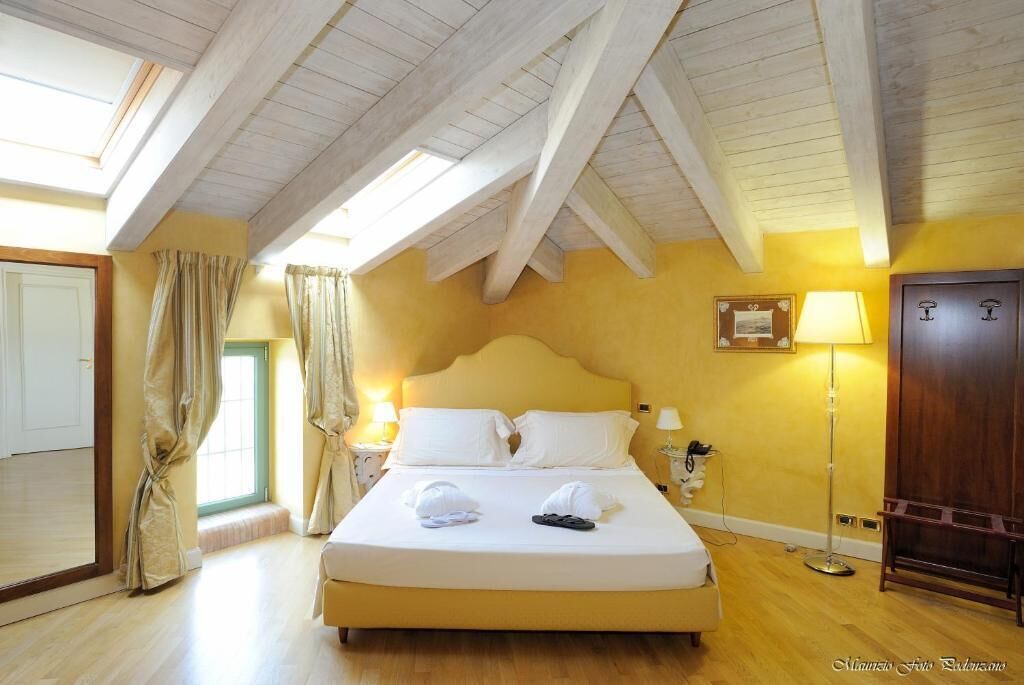 A hotel room with a high ceiling, a large bed, a bedside table with a lamp, and a wardrobe.
