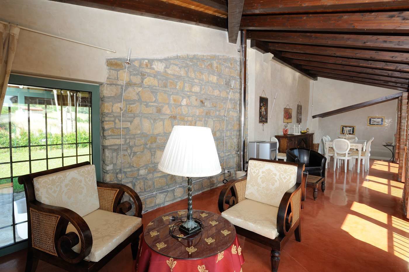 A cozy room with two upholstered chairs, a table with a lamp, a stone wall, and a dining area with white chairs.
