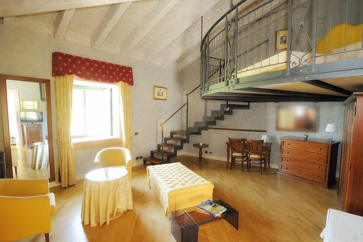 A spacious room with a wooden floor, a bed, a sofa, a table, a chair, a staircase, and a TV.