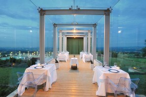 An outdoor dining area with tables set for a meal, a green curtain backdrop, and a view of the landscape. - Relais La Colombara (Travo)
