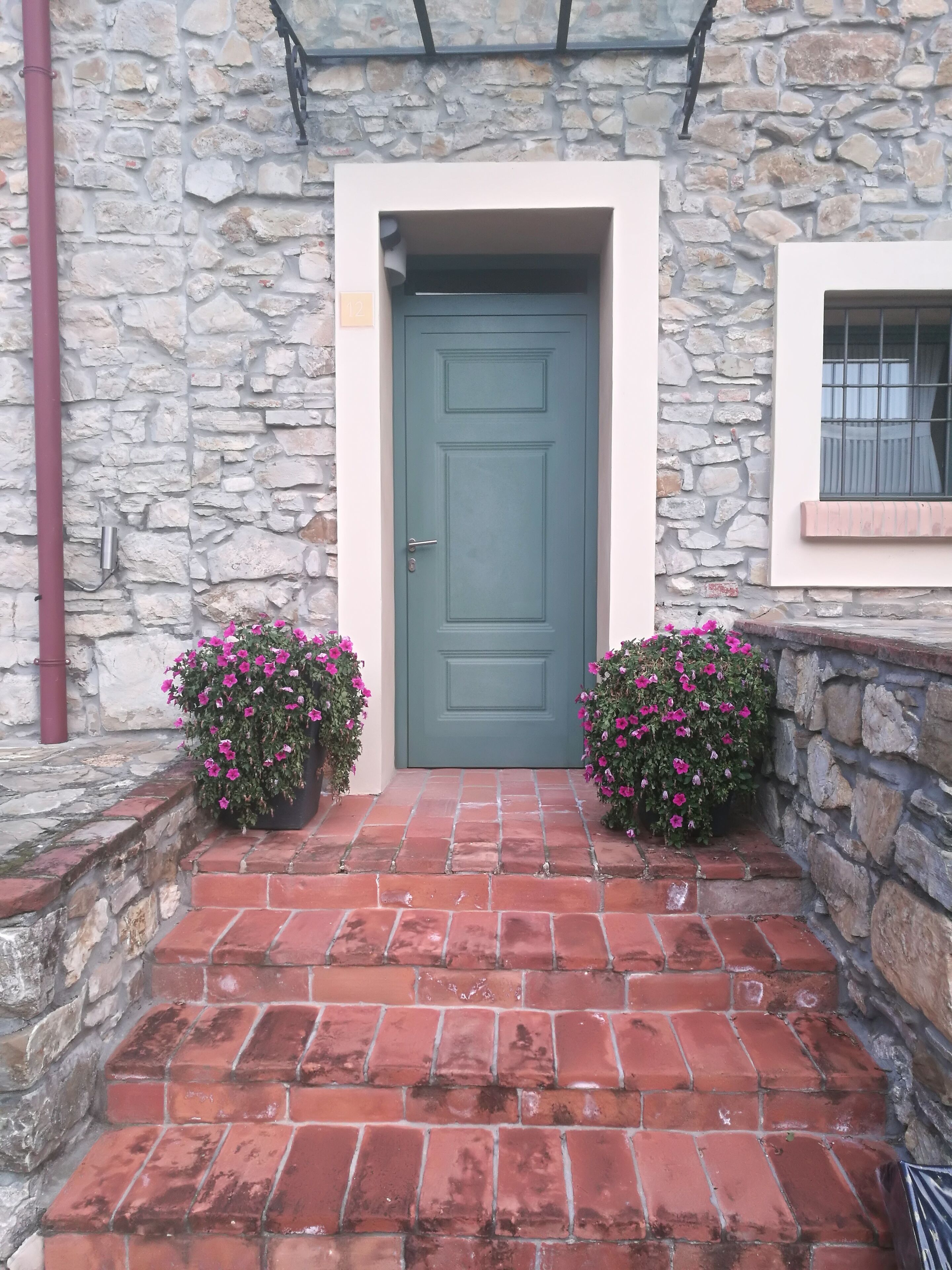 Un edificio de piedra con una puerta verde, dos macetas con flores rosadas y un camino de ladrillo.