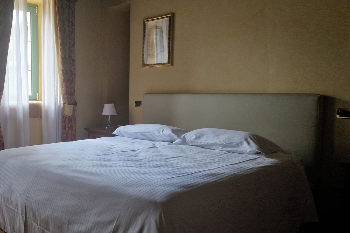 A neatly made bed in a hotel room with a large window, curtains, a bedside lamp, and a framed picture on the wall.