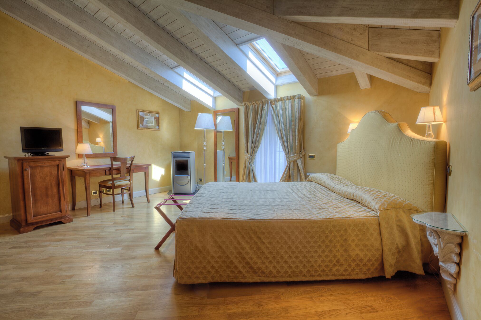 Une chambre à coucher avec un lit, un bureau avec un ordinateur, une télévision et une fenêtre avec des rideaux.