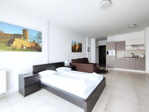 Quarto de hotel moderno com uma cama grande, uma escrivaninha, uma cadeira e uma cozinha compacta.