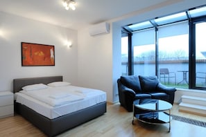 A modern bedroom with a bed, a black leather sofa, a small round table, and a large window with a view of the outdoors.