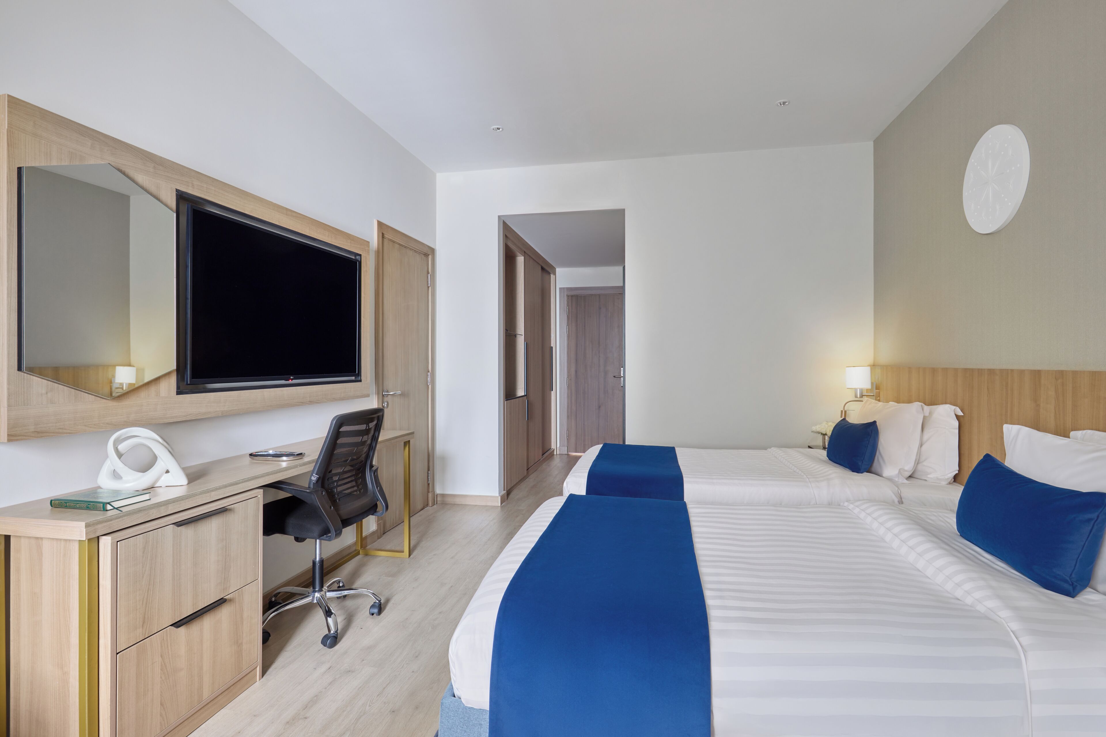 A hotel room with a desk, chair, two beds with blue bedding, a flat-screen TV, and a wall clock.