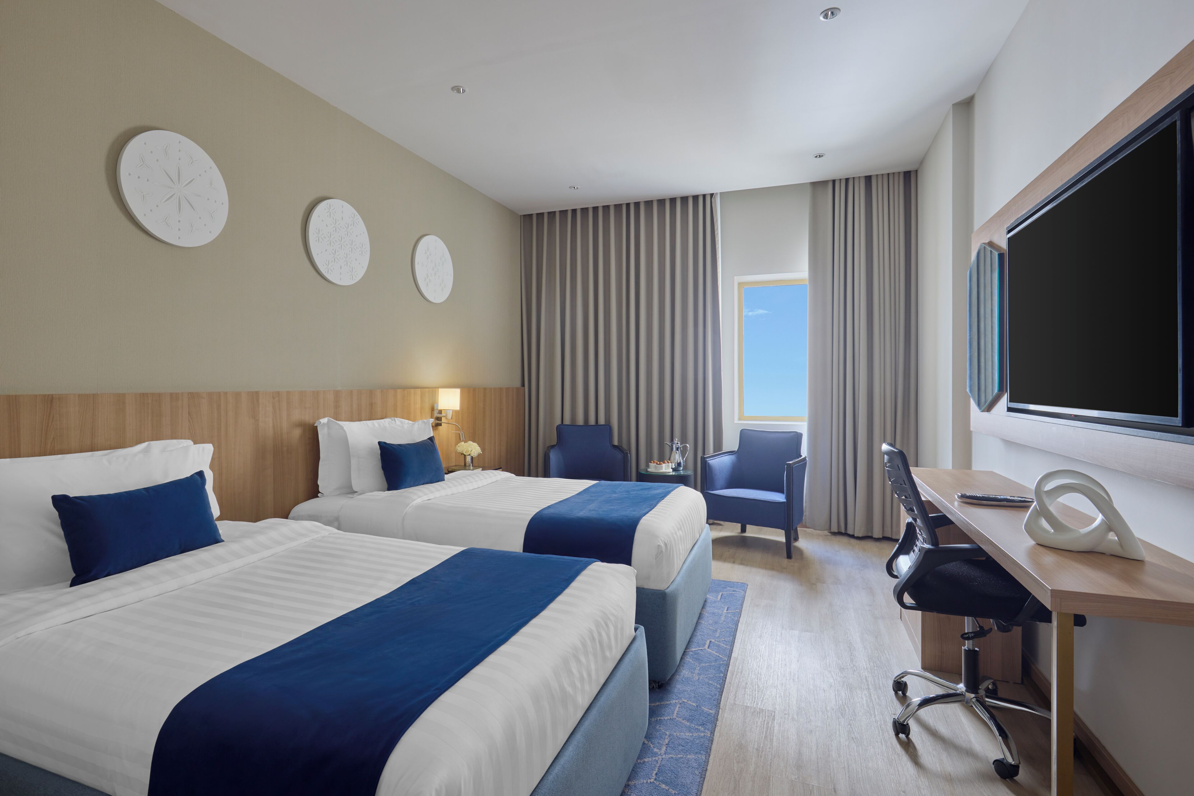 A hotel room with two beds, a desk, a chair, a TV, and a window with curtains.