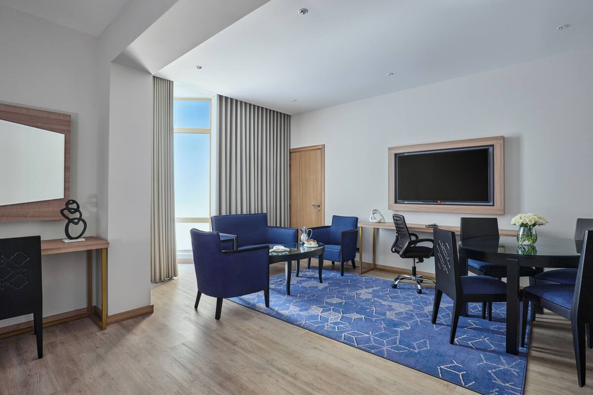 Een moderne hotelkamer met een grote flatscreen-tv, een zithoek met blauwe stoelen, een houten bureau en een groot raam met gordijnen.