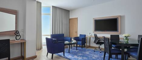 A modern hotel room with a large flat-screen TV, a seating area with blue chairs, a wooden desk, and a large window with curtains.