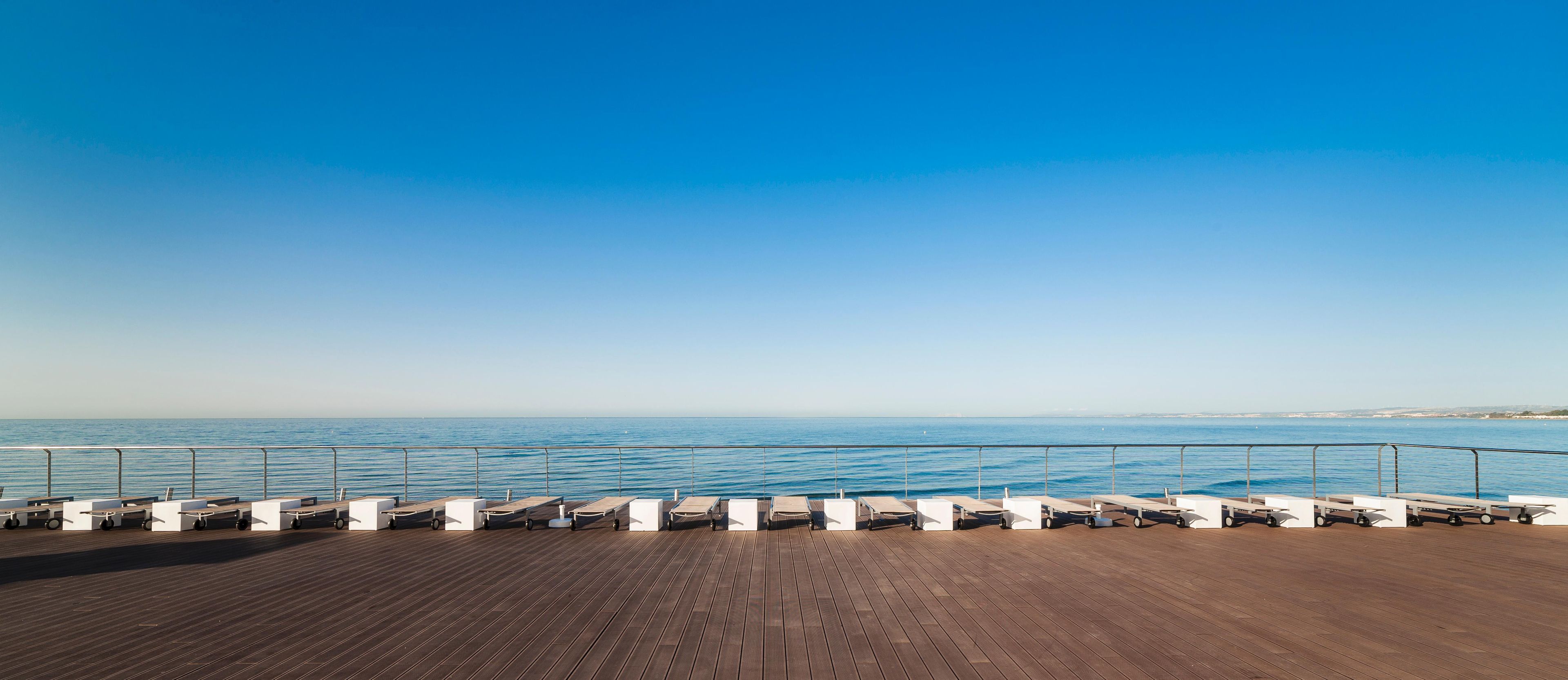 A wooden deck with sun loungers facing a calm sea under a clear sky.