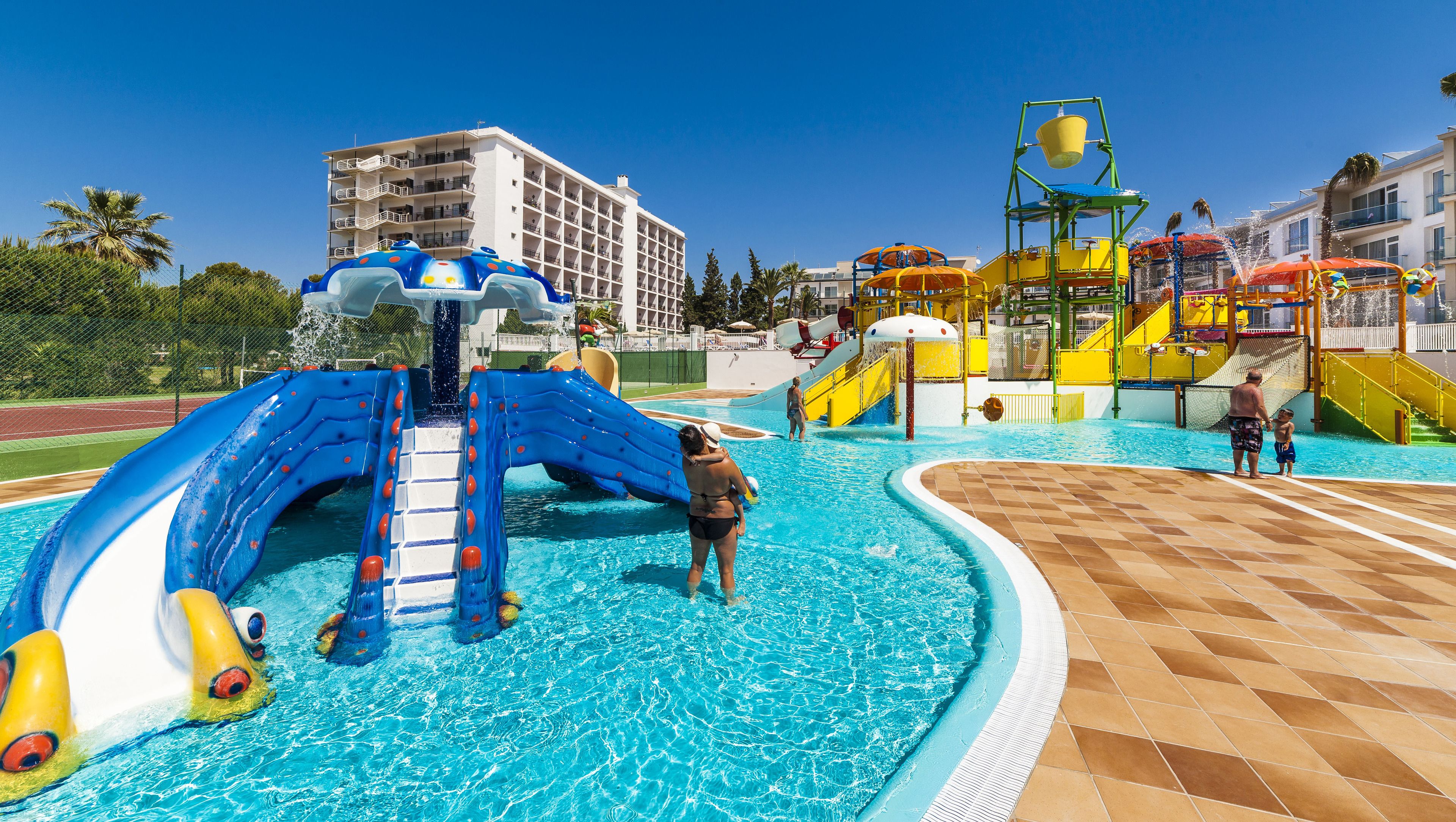 Parque acuático con toboganes y estructuras de juego, piscina y un edificio al fondo.