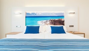 A double bed with a striped blue and white bedspread, two blue pillows, and a large framed picture of a coastal landscape on the wall behind. - Globales Playa Estepona (Estepona)