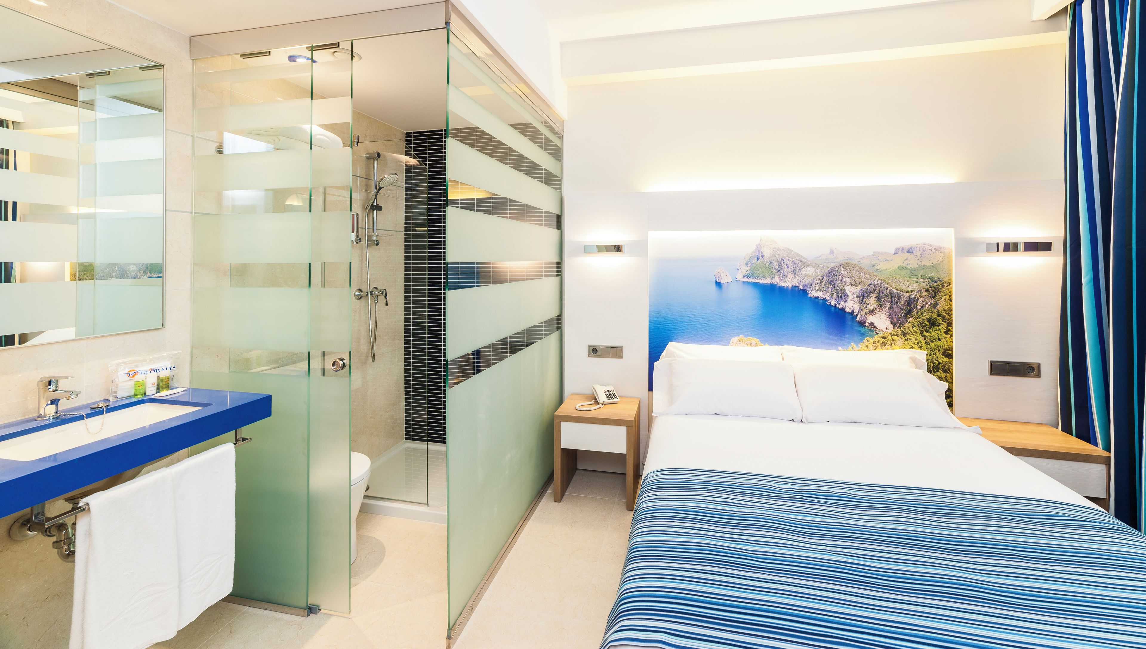 Een moderne hotelkamer met een douche in een glazen cabine, een groot bed met uitzicht op zee, een blauw gestreepte sprei en een badkamer met een blauw aanrechtblad en witte handdoeken.