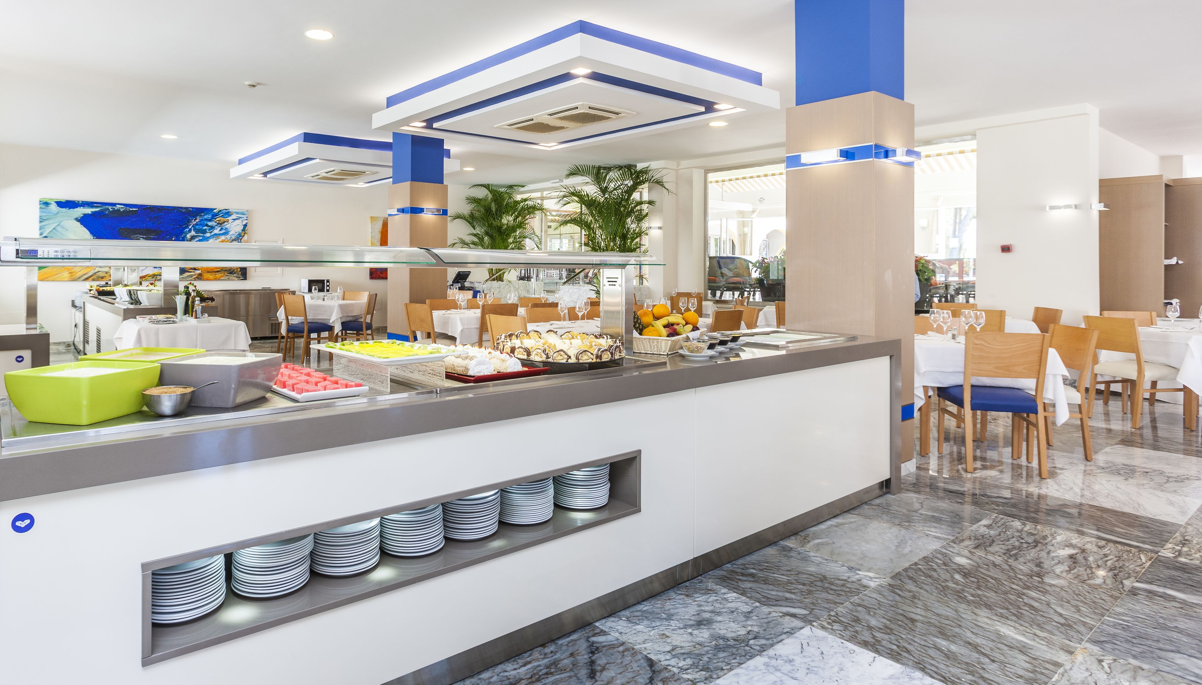 Uma área de buffet moderna com diversas opções de comida, incluindo frutas e sobremesas, e um espaço de estar com mesas e cadeiras.