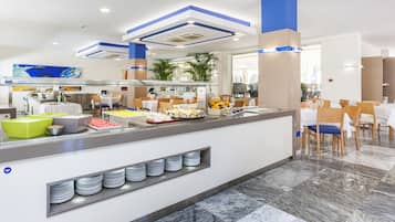 Uma área de buffet moderna com diversas opções de comida, incluindo frutas e sobremesas, e um espaço de estar com mesas e cadeiras.