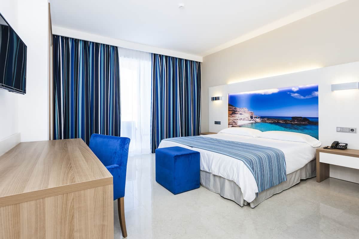 A hotel room with a bed, a desk, a chair, a phone, and a painting of a coastal scene.