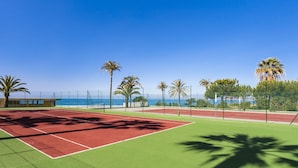A tennis court with a red and green surface, surrounded by palm trees and a fence, with a view of the ocean in the background. - Globales Playa Estepona (Estepona)