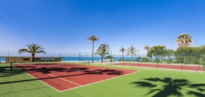 A tennis court with a red and green surface, surrounded by palm trees and a fence, with a view of the ocean in the background. - Globales Playa Estepona (Estepona)