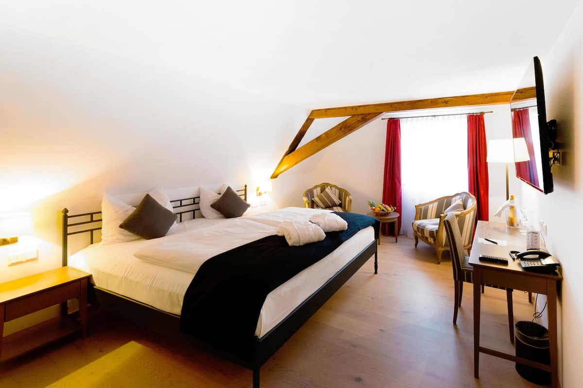 A bedroom with a bed, bedside table, chair, desk, lamp, and a window with curtains.