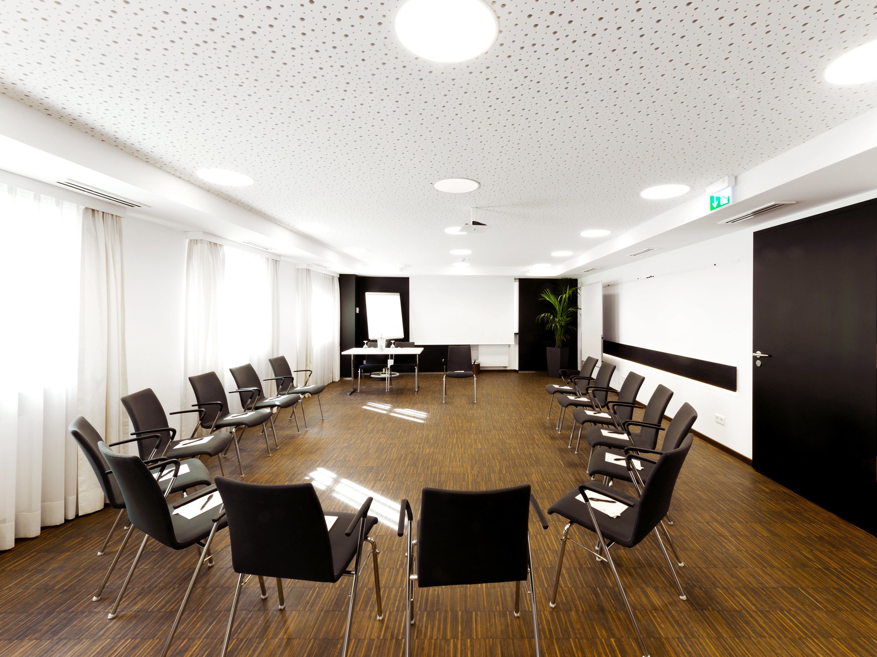 A conference room with a round table, black chairs, a projector screen, and a potted plant.