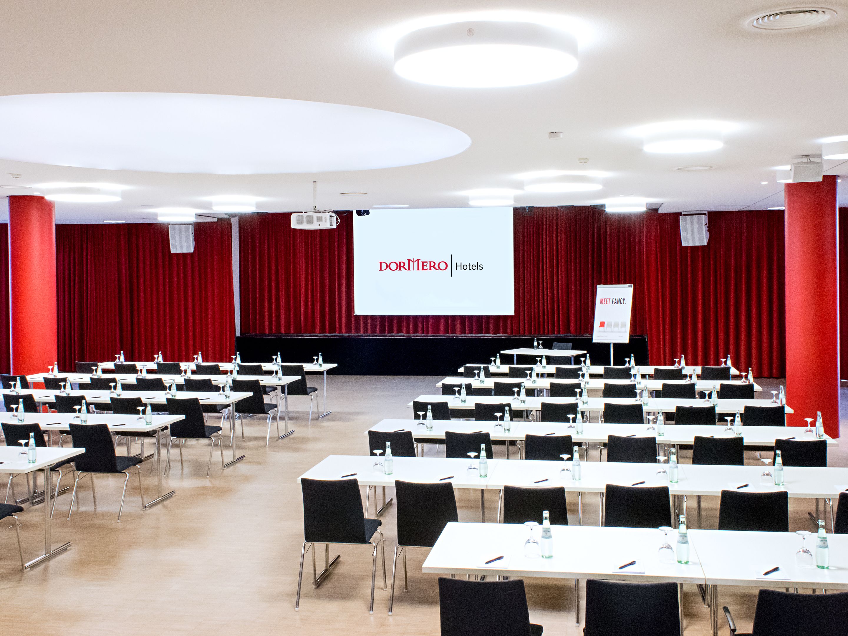 Un salón de conferencias con filas de mesas y sillas, una pantalla grande mostrando 'DORMERO Hotels' y un letrero que dice 'Meet Fancy'.