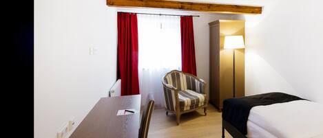 Una habitación de hotel pequeña con un escritorio de madera, un sillón a rayas, una cama individual y una lámpara de pie.
