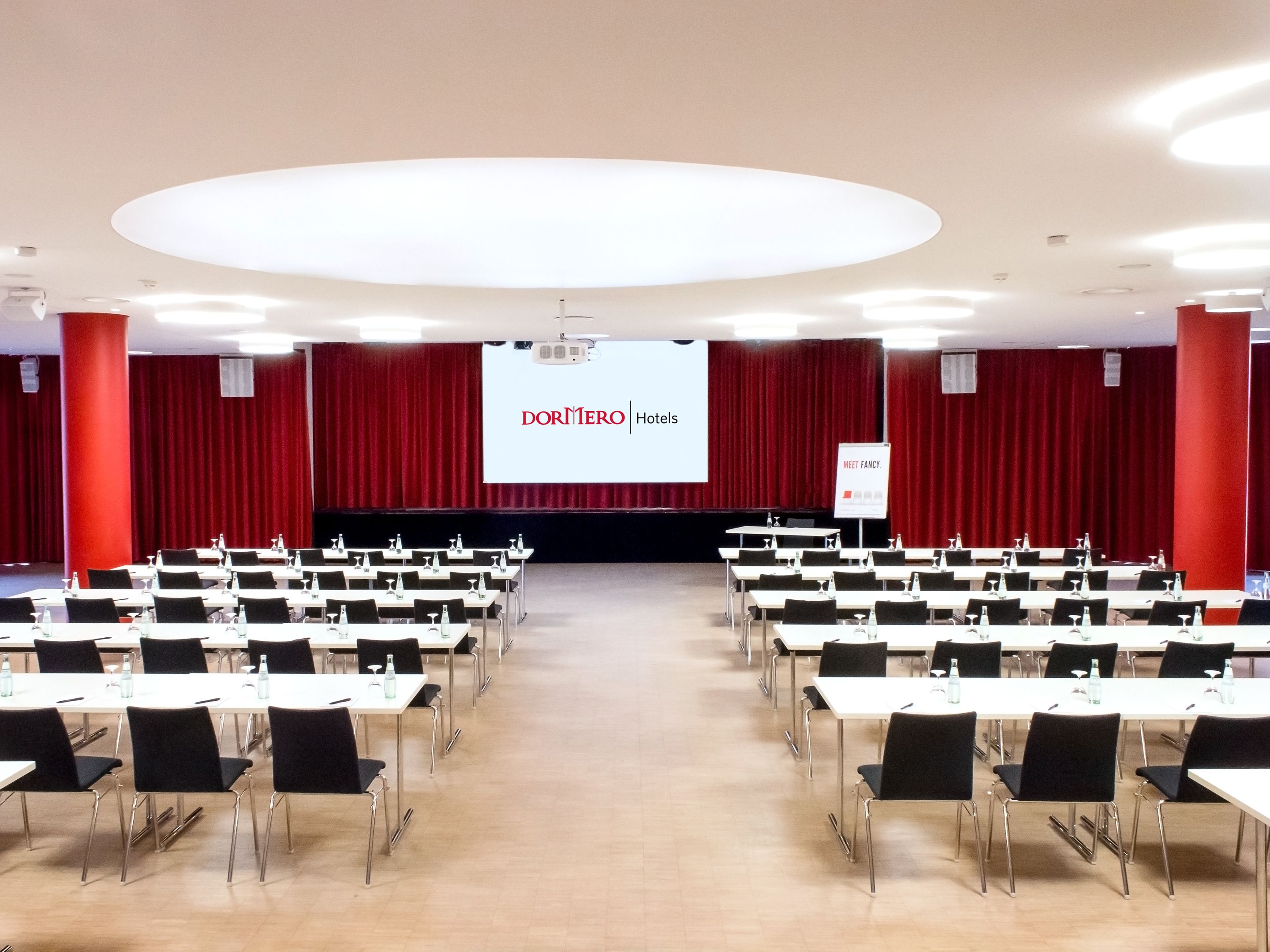 A conference room with rows of tables and chairs, a large screen at the front, and red curtains.