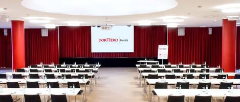 Un salón de conferencias con filas de mesas y sillas, una pantalla grande al frente y cortinas rojas.