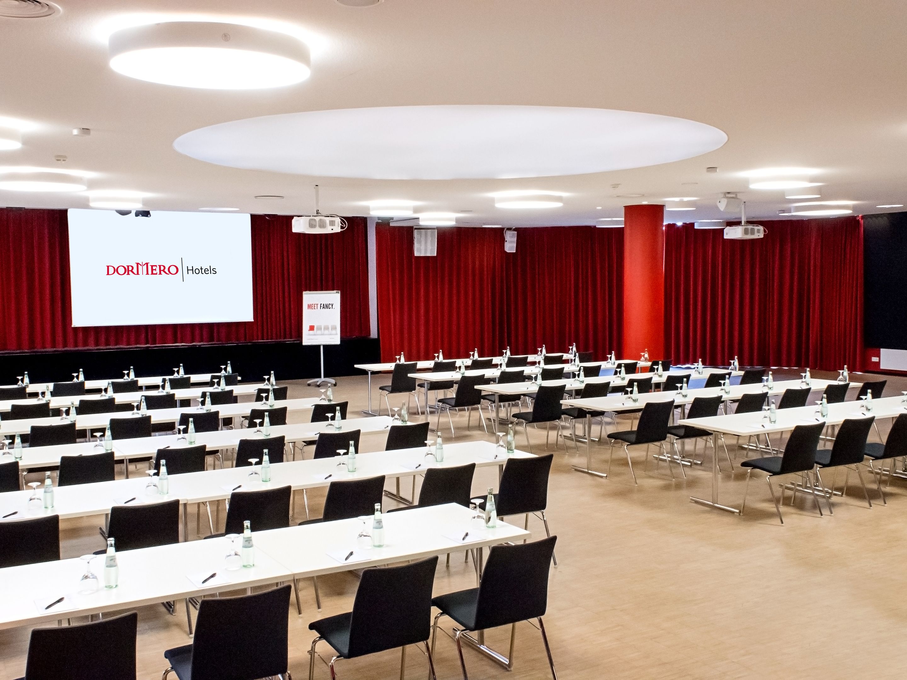 A conference room with rows of tables and chairs, a large screen displaying 'DORNER HOTEL', and a 'MEET FANCY' sign.