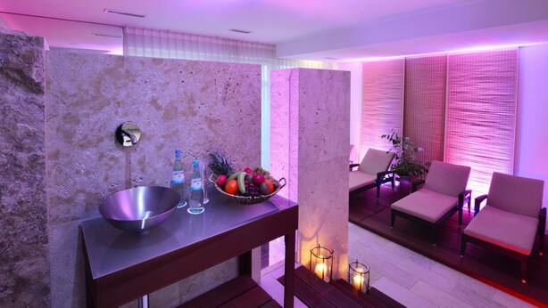 Un área de spa moderna con pared de piedra, un lavabo de metal, botellas y un frutero. Hay dos sillas y una mesa con velas.