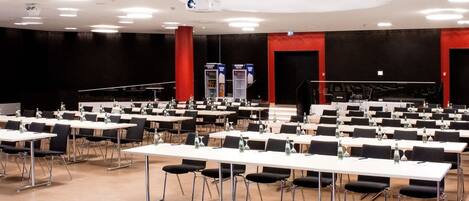 A conference room with multiple tables and chairs, a stage with a podium, and a projector on the ceiling.