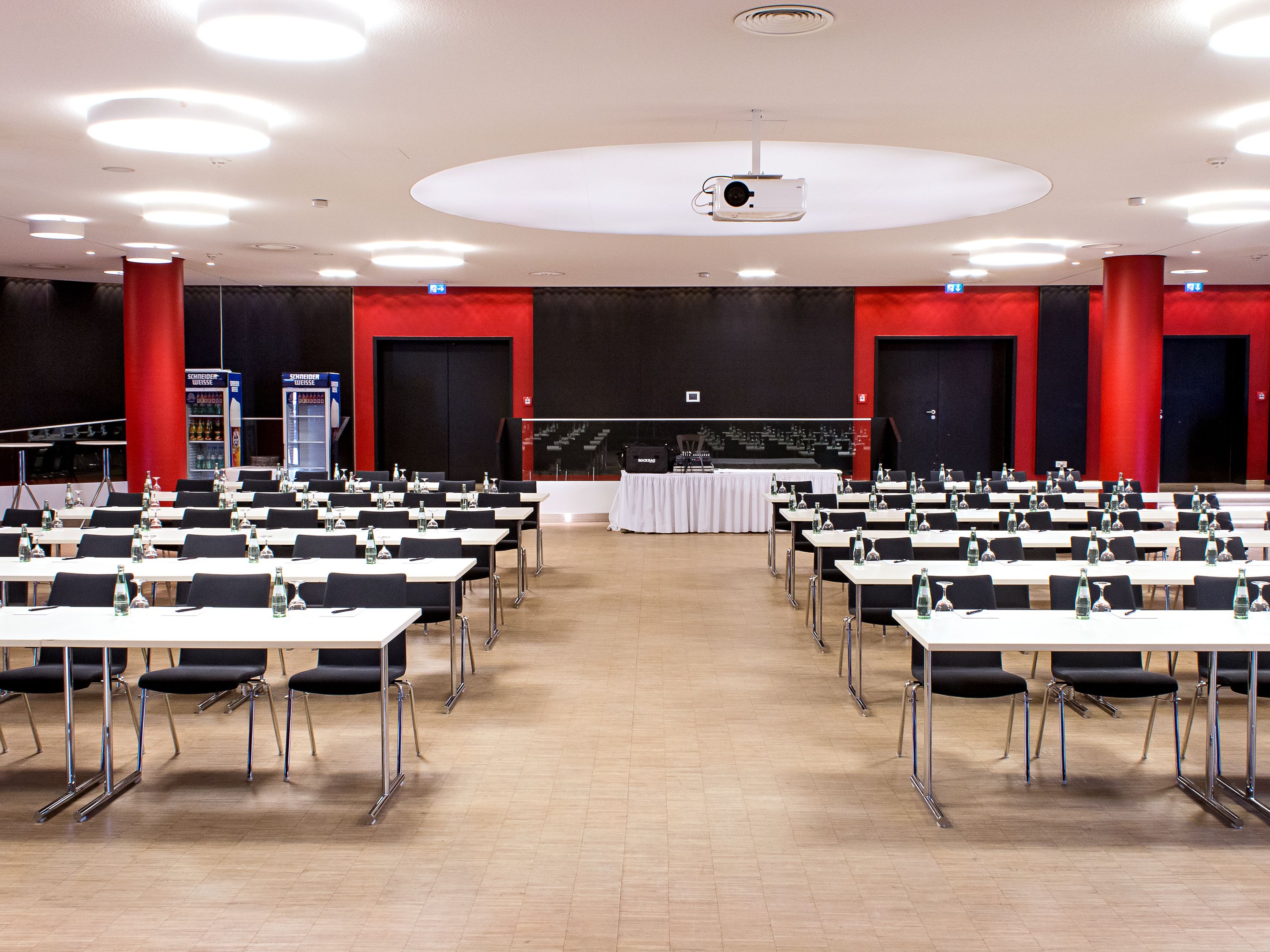 Un salón de conferencias con filas de mesas y sillas, un proyector en el techo y un escenario con un telón de fondo rojo.