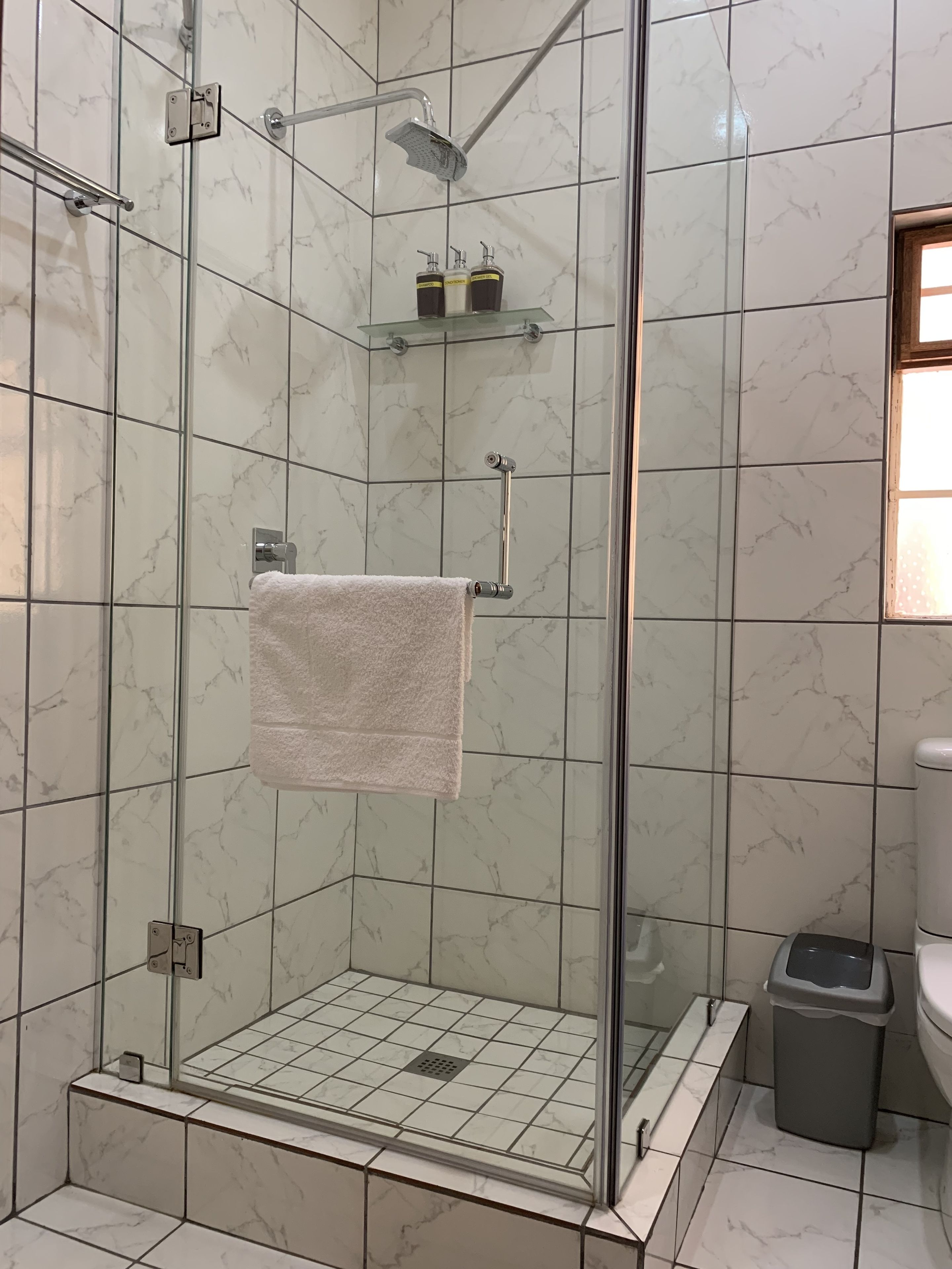 Ducha con cerramiento de vidrio, pared de azulejo de mármol blanco, repisa de vidrio con artículos de aseo y toalla blanca en un perchero.